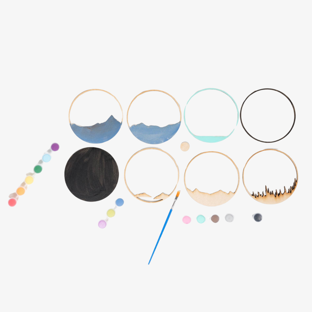 A Paint By Layers Craft Kit featuring a circular shadow box with a pre-designed landscape scene. The scene depicts a mountain range under a starry night sky, with a river flowing through a valley. The kit includes layers of wood, paint, and instructions. The title of the kit is "Adventure Awaits Shadow Box."
