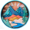 A circular layered wooden art piece depicting a mountain landscape with a sunset, snow, trees, and a winding river, shown on a white background.