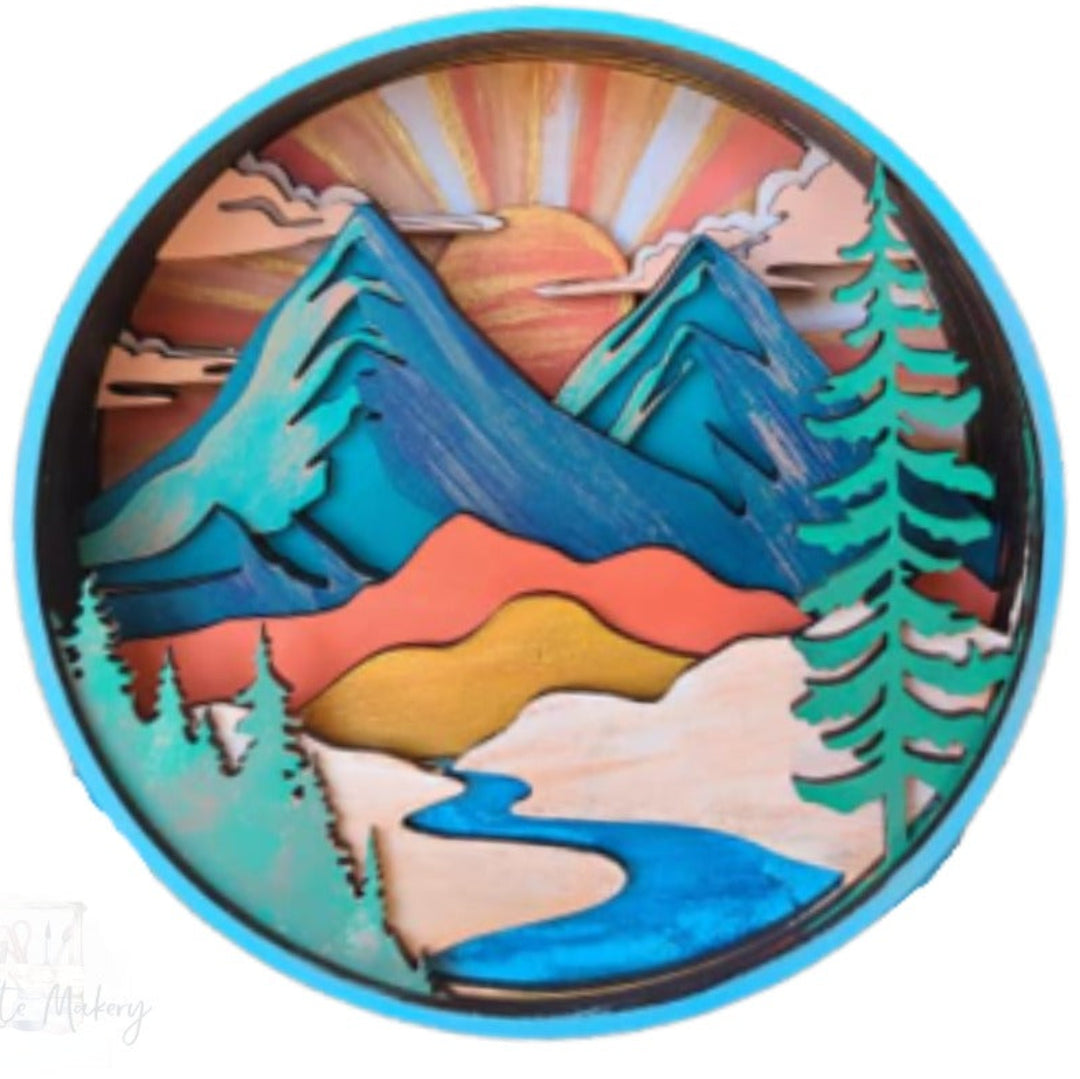 A circular layered wooden art piece depicting a mountain landscape with a sunset, snow, trees, and a winding river, shown on a white background.