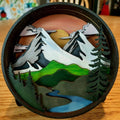 A Paint By Layers Craft Kit featuring a circular shadow box with a pre-designed landscape scene. The scene depicts a mountain range under a starry night sky, with a river flowing through a valley. The kit includes layers of wood, paint, and instructions. The title of the kit is "Adventure Awaits Shadow Box."