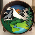 A Paint By Layers Craft Kit featuring a circular shadow box with a pre-designed landscape scene. The scene depicts a mountain range under a starry night sky, with a river flowing through a valley. The kit includes layers of wood, paint, and instructions. The title of the kit is "Adventure Awaits Shadow Box."