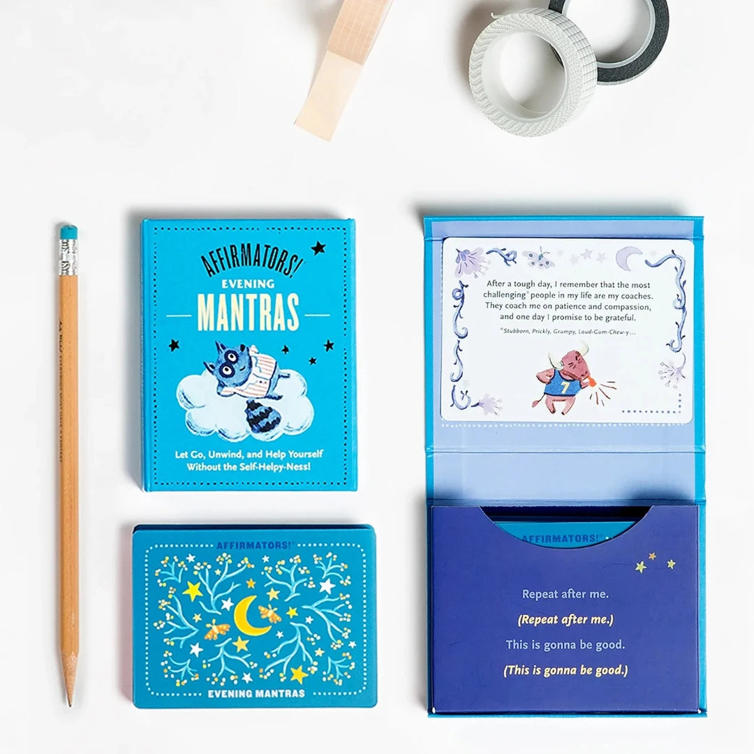 A pink box containing a deck of affirmation cards. The box has the text "AFFIRMATORS! EVENING MANTRAS" on the front. The cards have various affirmations printed on them, including "Let Go, Unwind, and Help Yourself Without the Self-Helpy-Ness!" and "Thank you to me! For always being willing to try, what's next." The box also includes a small booklet with additional affirmations.