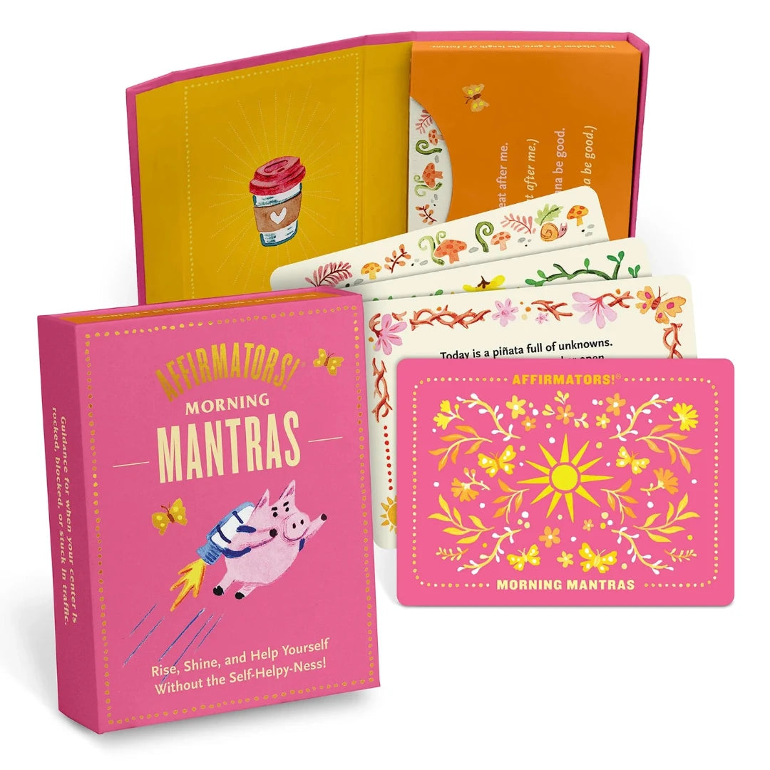 A pink box containing a deck of affirmation cards. The box has the text "AFFIRMATORS! MORNING MANTRAS" on the front. The cards have various affirmations printed on them, including "Rise, Shine, and Help Yourself Without the Self-Helpy-Ness!" and "Today is a piñata full of unknowns." The box also includes a small booklet with additional affirmations.