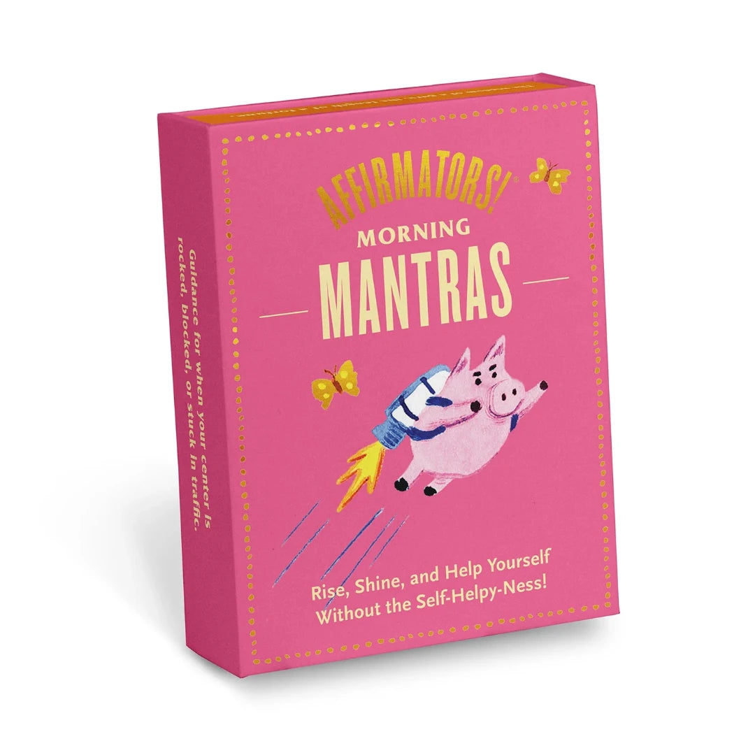 A pink box containing a deck of affirmation cards. The box has the text "AFFIRMATORS! MORNING MANTRAS" on the front. The cards have various affirmations printed on them, including "Rise, Shine, and Help Yourself Without the Self-Helpy-Ness!" and "Today is a piñata full of unknowns." The box also includes a small booklet with additional affirmations.