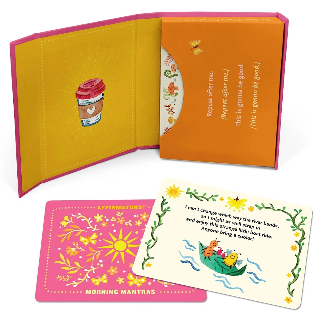 A pink box containing a deck of affirmation cards. The box has the text "AFFIRMATORS! MORNING MANTRAS" on the front. The cards have various affirmations printed on them, including "Rise, Shine, and Help Yourself Without the Self-Helpy-Ness!" and "Today is a piñata full of unknowns." The box also includes a small booklet with additional affirmations.