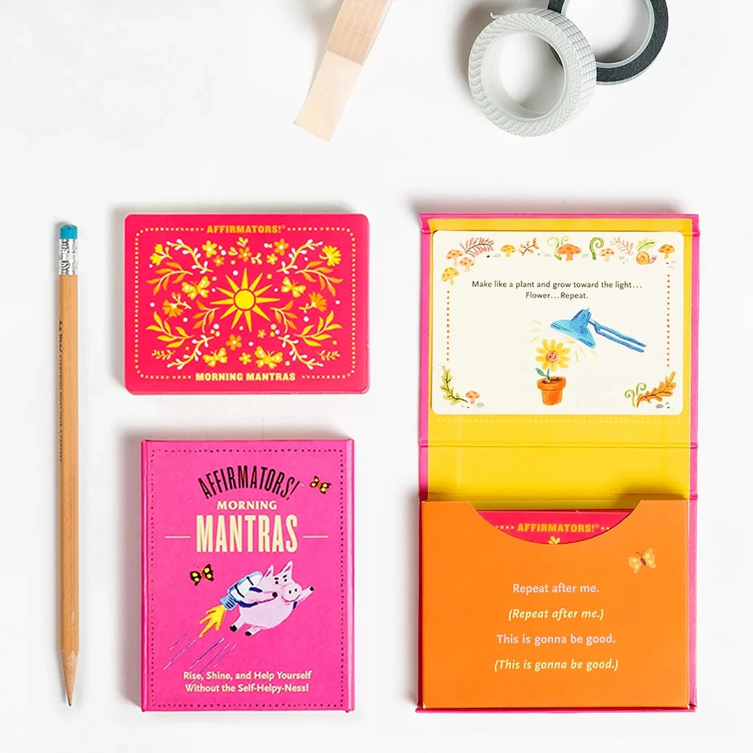 A pink box containing a deck of affirmation cards. The box has the text "AFFIRMATORS! MORNING MANTRAS" on the front. The cards have various affirmations printed on them, including "Rise, Shine, and Help Yourself Without the Self-Helpy-Ness!" and "Today is a piñata full of unknowns." The box also includes a small booklet with additional affirmations.