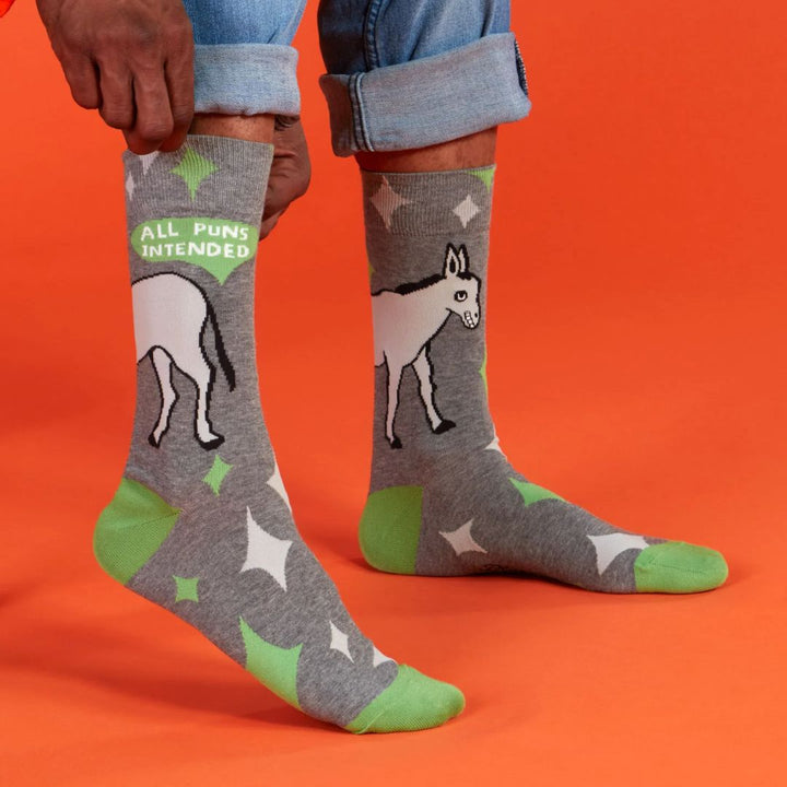 A pair of men's crew socks with a grey background and a pattern of white starbursts and green accents. One sock features the rear end of a white donkey and the text 