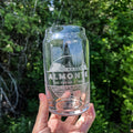 A clear glass with etched designs of a can and text that reads 'Almonte The Friendly Town', held in front of a tree backdrop.