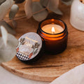 Candle in a glass jar with a decorative label on a wooden surface, surrounded by eucalyptus leaves.