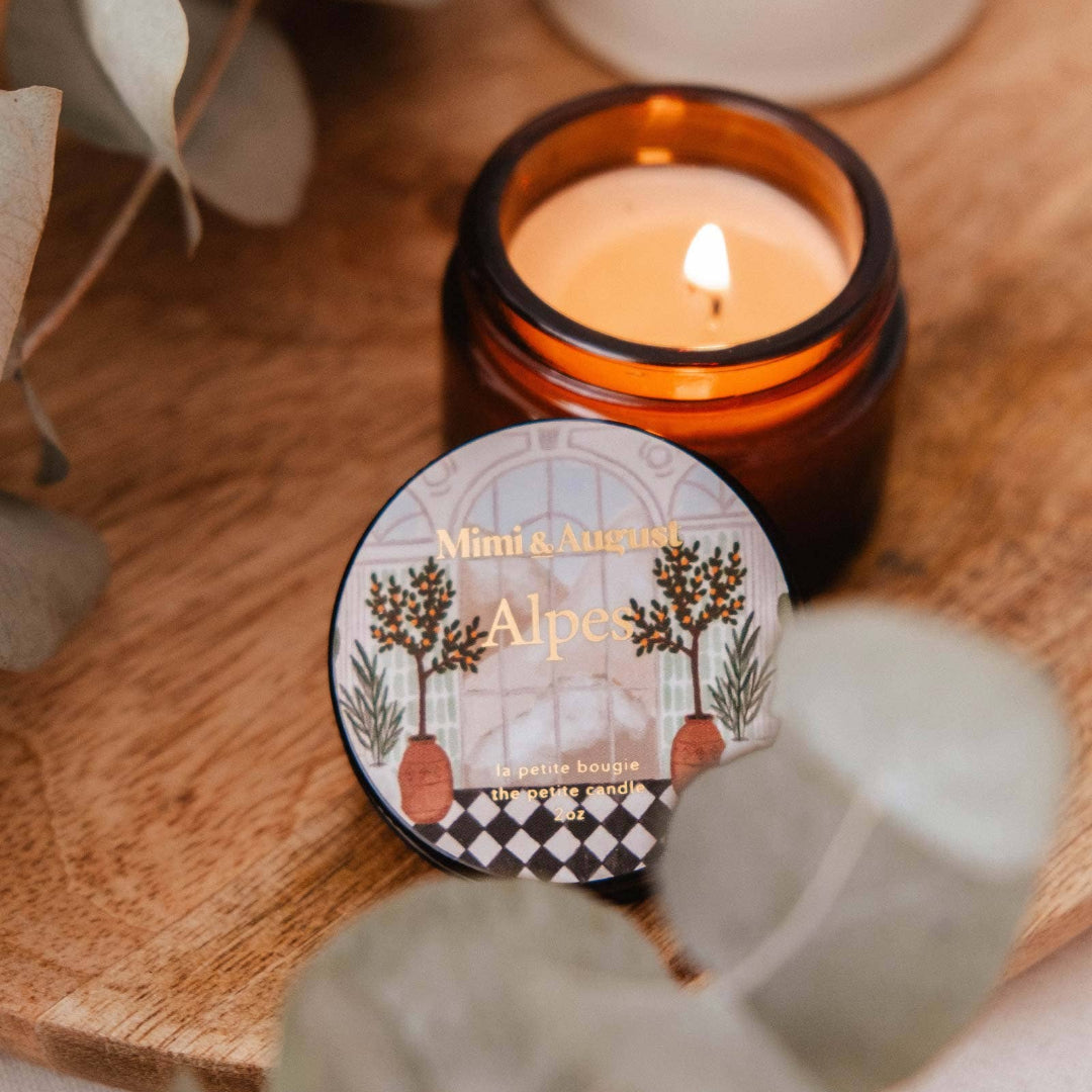 Candle in an amber jar with a decorative label on a wooden surface, surrounded by eucalyptus leaves.
