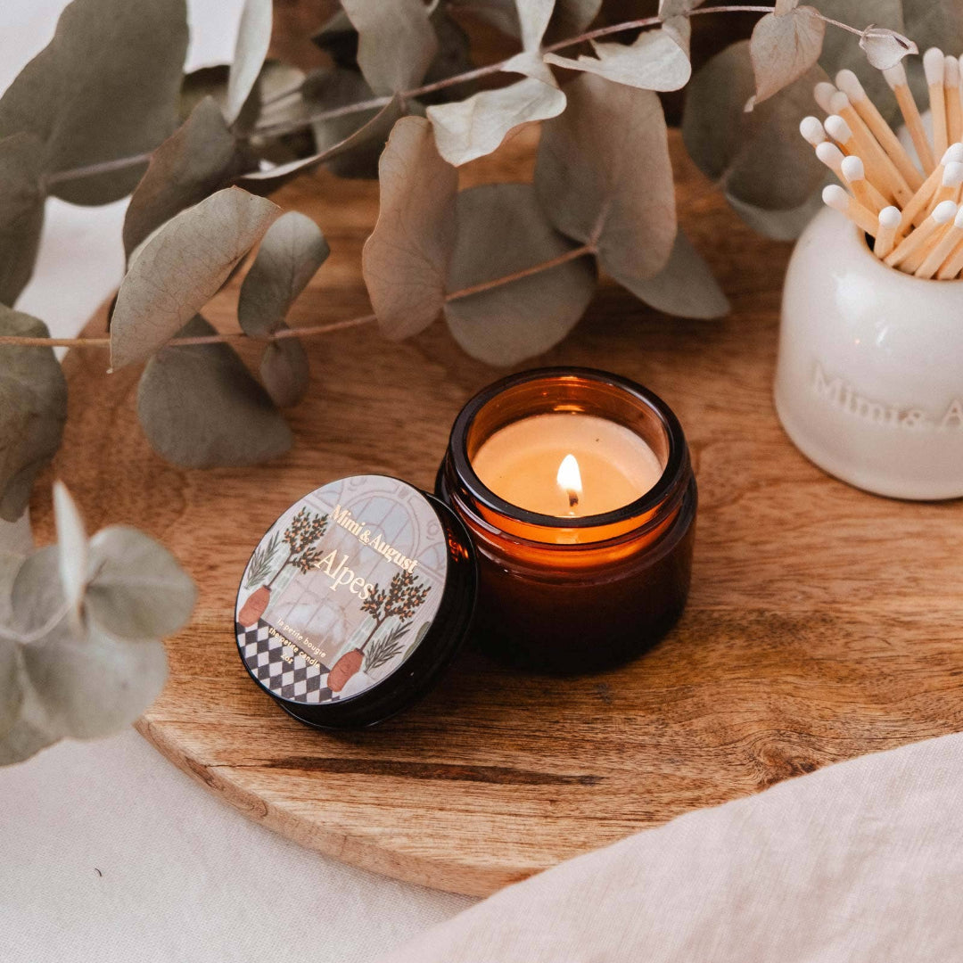Small lit candle in a black holder on a wooden surface with eucalyptus leaves and a matchbox.