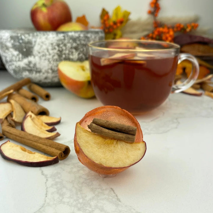 An orange, spherical tea bomb topped with a cinnamon stick and apple slices. The tea bomb is resting on a white marble surface.