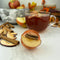 An orange, spherical tea bomb topped with a cinnamon stick and apple slices. The tea bomb is resting on a white marble surface.