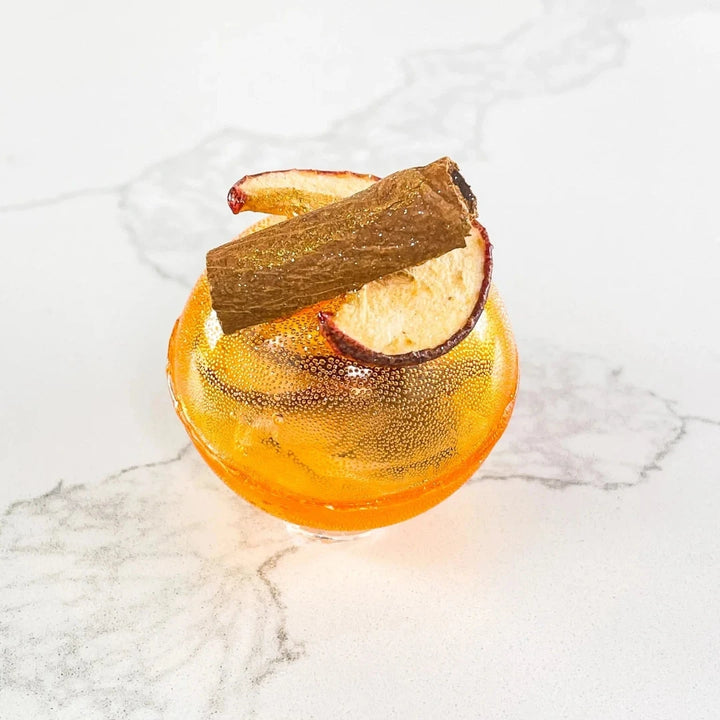 An orange, spherical tea bomb topped with a cinnamon stick and apple slices. The tea bomb is resting on a white marble surface.