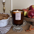 Lit candle in a brown glass jar labeled 'apple crumble' on a decorative stand with apples and cinnamon sticks in the background.