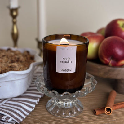 Lit candle in a brown glass jar labeled 'apple crumble' on a decorative stand with apples and cinnamon sticks in the background.