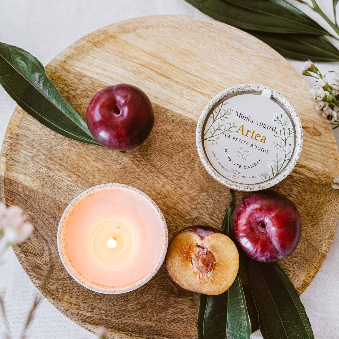 Candle, plums, and a container labeled 'Artea' on a wooden board with leaves.