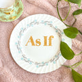 Decorative plate with 'As If' text on a pink surface with green leaves