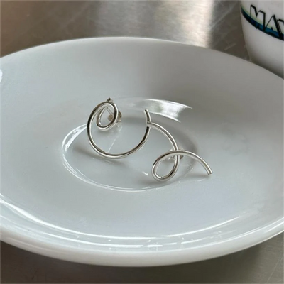 A pair of asymmetric twist earrings made of sterling silver, featuring a unique, abstract, and mismatched swirling design, displayed on a white plate. These modern earrings are available in Ottawa, Ontario, Canada