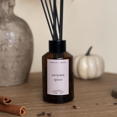 Diffuser bottle labeled 'autumn spice' by North + Rose on a wooden surface with decorative elements.