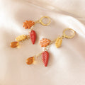 A pair of gold-tone hoop earrings with dangling autumn leaf charms in orange, yellow, and red, displayed on a soft beige satin fabric background.
