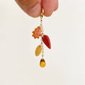 A close-up of a hand holding a gold-tone keychain with dangling autumn leaf charms in orange, red, and yellow, and a small amber teardrop bead at the bottom, against a plain off-white background.