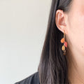 A close-up side view of a person wearing a dangling earring with autumn leaf charms in orange, red, and gold, along with a small amber bead drop, against a plain light wall.