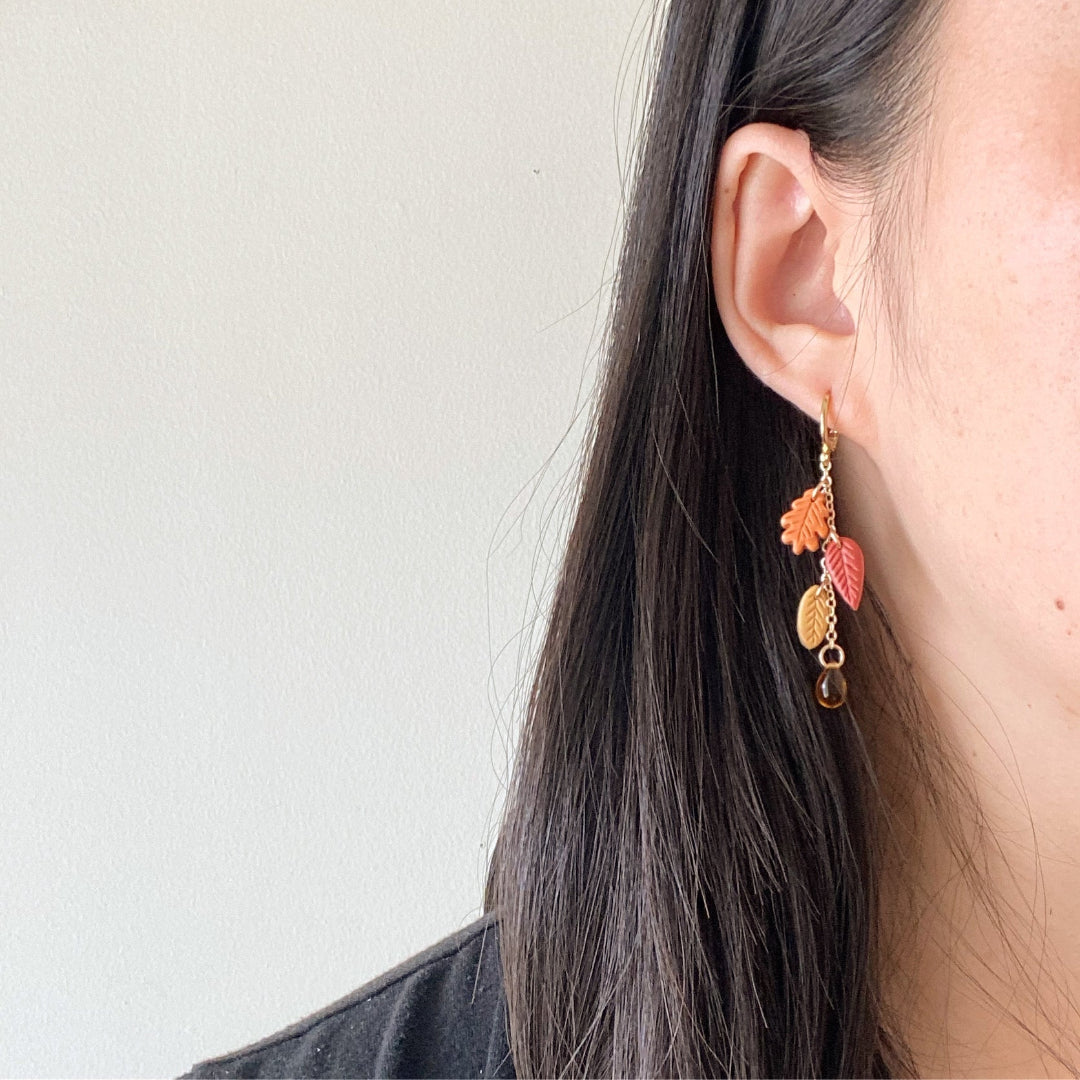 A close-up side view of a person wearing a dangling earring with autumn leaf charms in orange, red, and gold, along with a small amber bead drop, against a plain light wall.