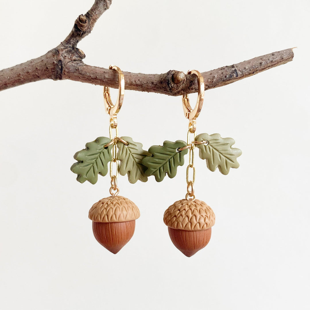 Acorn and leaf drop earrings hanging from a small branch against a clean white background. Each earring has a gold hoop, green leaf charms, and a wooden acorn charm with a tan textured cap and brown acorn body.