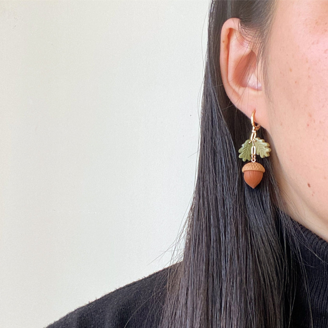 A close-up of a person wearing a dangling earring shaped like an acorn with a green leaf above it, against a plain light background.