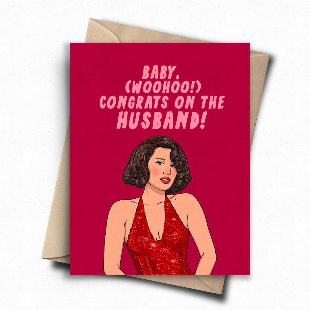 A greeting card with a kraft envelope, featuring a bright pink-red background with the text 'BABY, (WOOOH!) CONGRATS ON THE HUSBAND!' above an illustrated woman in a red sequined dress.