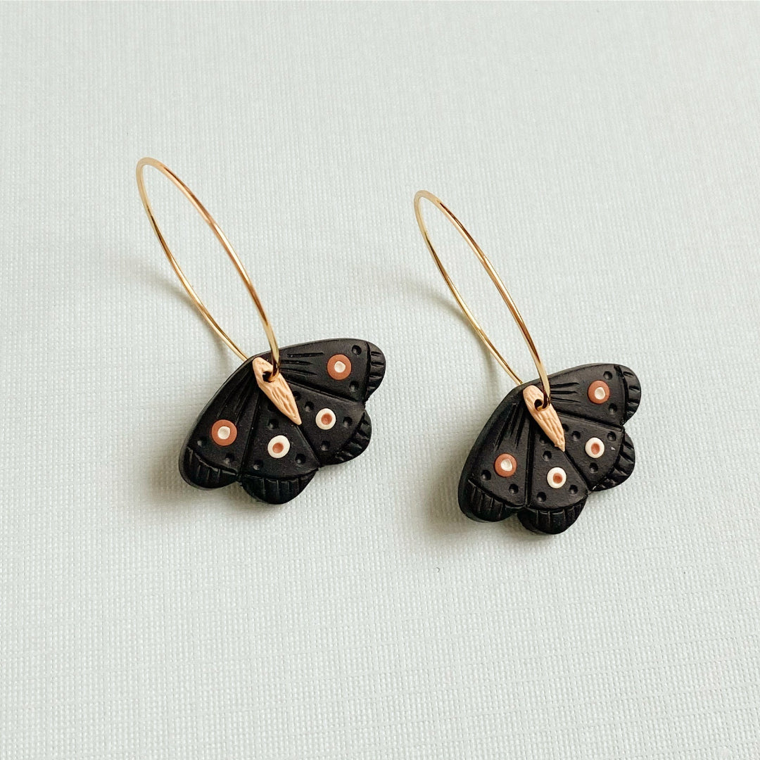 A pair of black butterfly earrings with gold-tone wire loops and small orange, white, and black eye details, displayed on a light textured background.