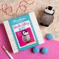 Needle felting kit with felted penguin, balls of yarn, and a needle on a wooden surface.