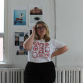Person wearing a white t-shirt with red graphics in a room with white walls and a window.