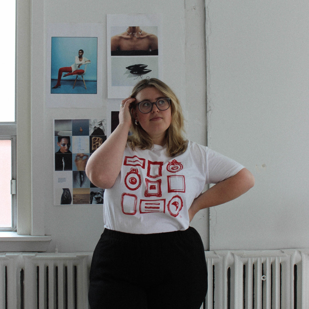 Person wearing a white t-shirt with red graphics in a room with white walls and a window.