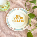 Decorative plate with 'Be Your Selfie' text on a textured surface with leaves.