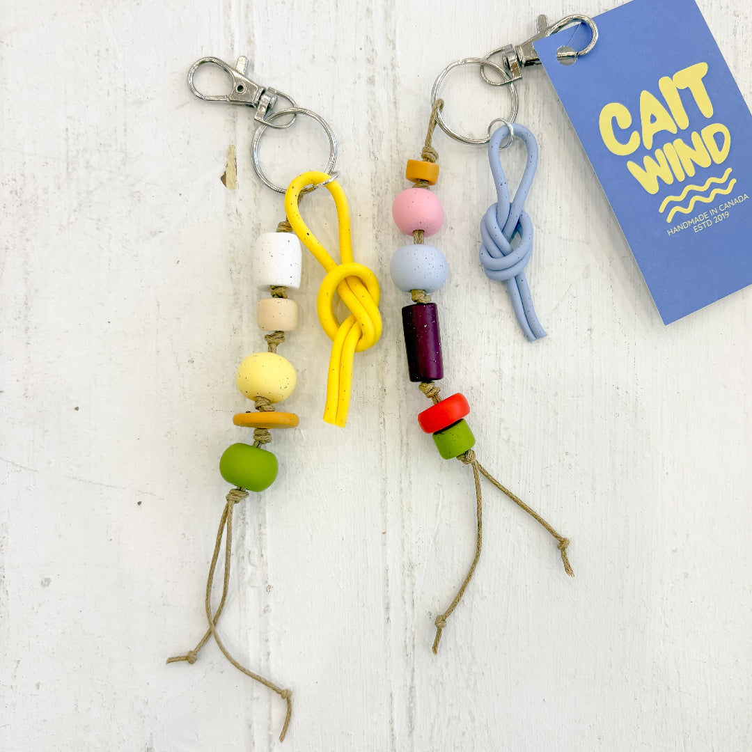 Colorful beaded keychains on a white wooden surface with a 'Cait Wind' branded card.