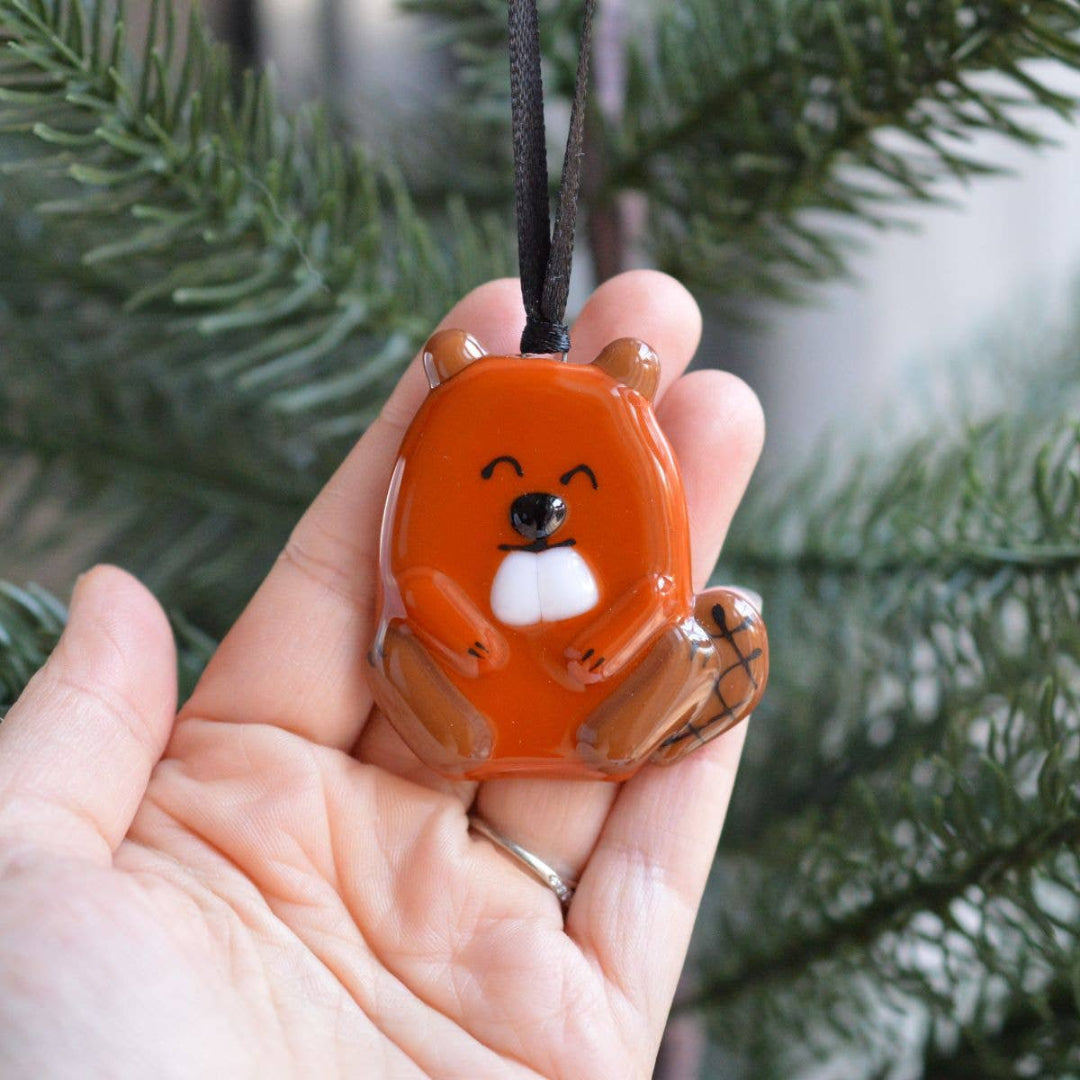A hand holding an orange beaver-shaped glass ornament with a black hanging ribbon, with blurred evergreen branches in the background.