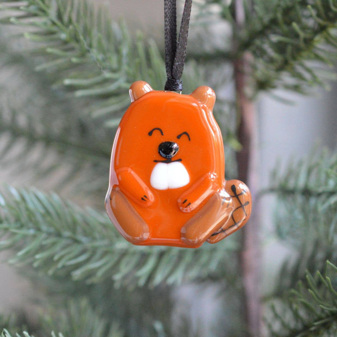 An orange beaver-shaped hanging ornament with a black hanging string, shown suspended in front of blurred evergreen branches.