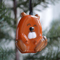 A glossy ceramic beaver ornament in shades of orange and brown with a white snout, hanging from a black ribbon against a blurred green and white background.