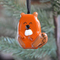 An orange beaver-shaped Christmas ornament with a white snout, black eyes, and black stitched details on its tail, hanging from a black string against a blurred green pine background.