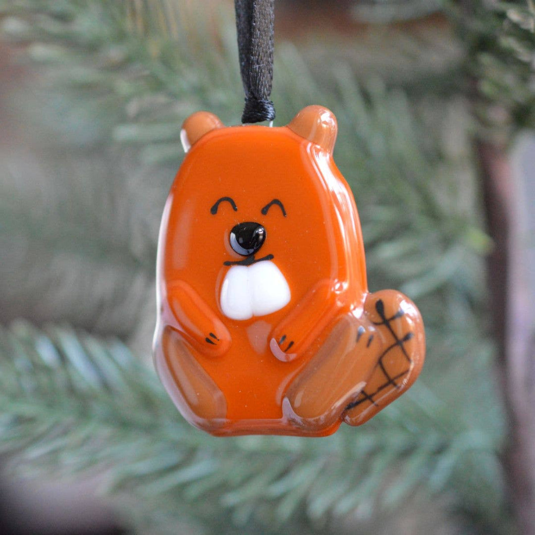 An orange beaver-shaped Christmas ornament with a white snout, black eyes, and black stitched details on its tail, hanging from a black string against a blurred green pine background.