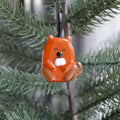 An orange beaver Christmas ornament hanging from a black ribbon on a green Christmas tree branch.