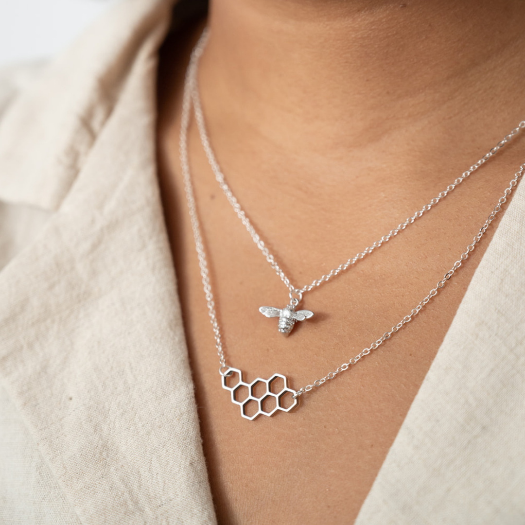 A pair of silver necklaces on a white card. The longer necklace features a honeycomb pendant, and the shorter necklace features a bee pendant.