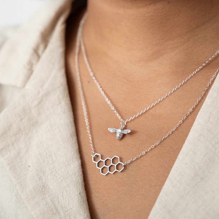 A pair of silver necklaces on a white card. The longer necklace features a honeycomb pendant, and the shorter necklace features a bee pendant.