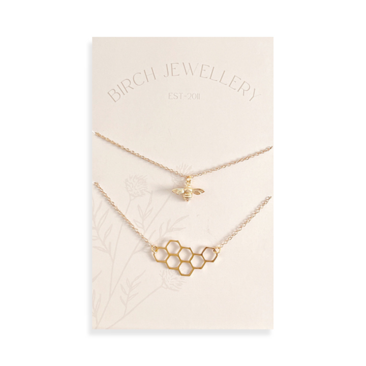 A pair of gold necklaces on a white card. The longer necklace features a honeycomb pendant, and the shorter necklace features a bee pendant. The card has the text 