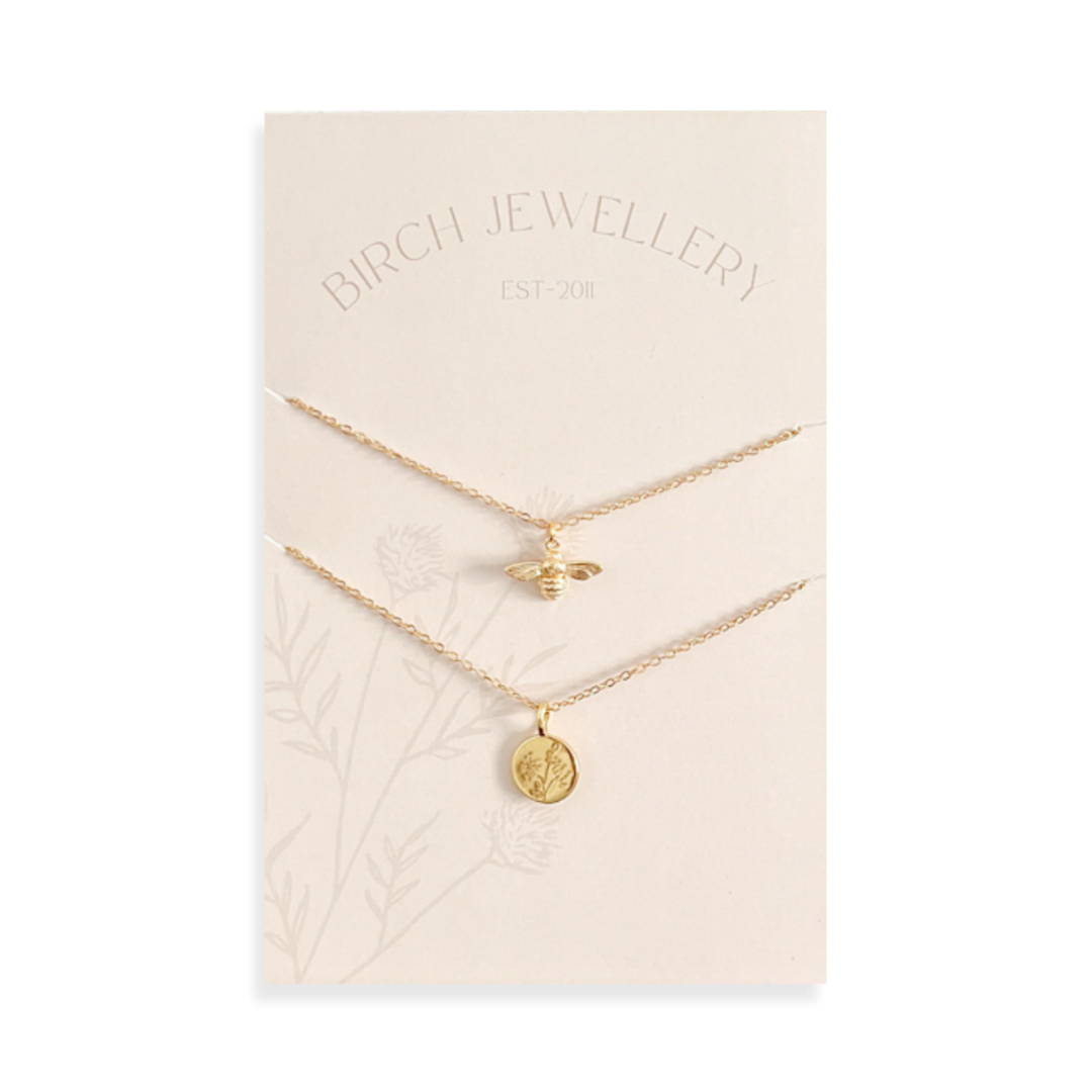 A pair of gold necklaces on a white card. The longer necklace features a honeycomb pendant, and the shorter necklace features a bee pendant. 