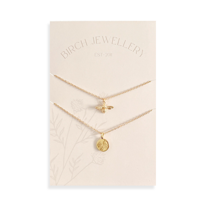 A pair of gold necklaces on a white card. The longer necklace features a honeycomb pendant, and the shorter necklace features a bee pendant. 