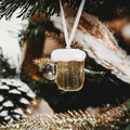 A miniature beer mug Christmas ornament with a white foamy head and a hanging white ribbon, shown hanging on a decorated Christmas tree.