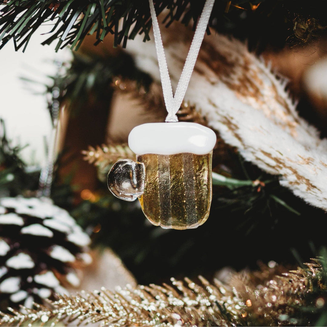 A miniature beer mug Christmas ornament with a white foamy head and a hanging white ribbon, shown hanging on a decorated Christmas tree.